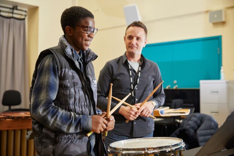 A boy in a blue checked t-shirt and camouflaged gilet plays the drums. He is stood next to a man in a grey shirt. Both are smiling.